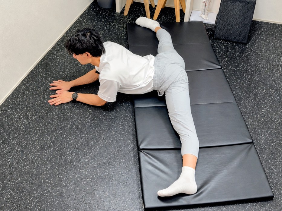 トレーナーが床で横開脚しながら上体を前に倒している